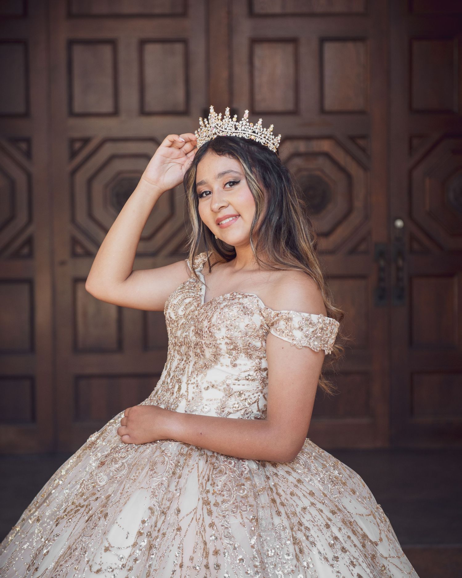 Young woman in gold quinceañera ball gown and tiara crown poses elegantly before ornate wooden doors.