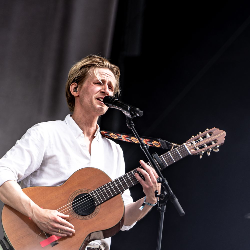 Male musician in white shirt playing acoustic guitar and singing into microphone on outdoor stage.