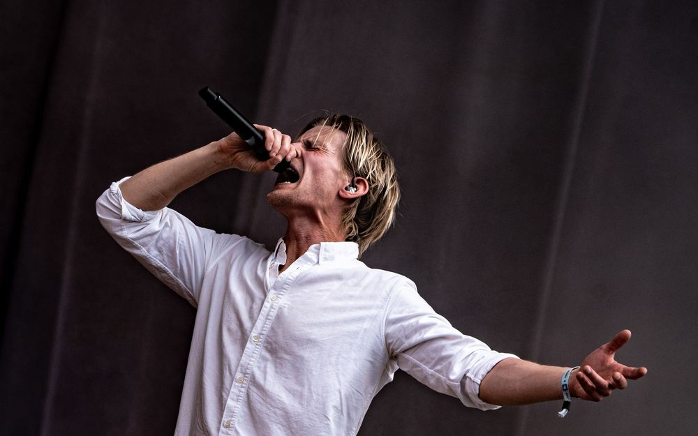 Male singer performing on stage, passionately singing into microphone wearing white shirt at outdoor concert.