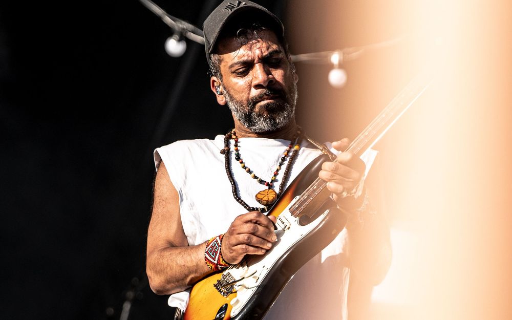 Bearded male guitarist performing on stage, wearing a white sleeveless shirt and cap, playing electric guitar with beaded necklace.