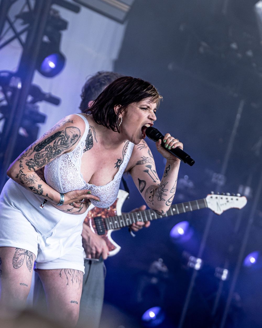 Tattooed female rock singer performing on stage, singing into microphone while playing white electric guitar.