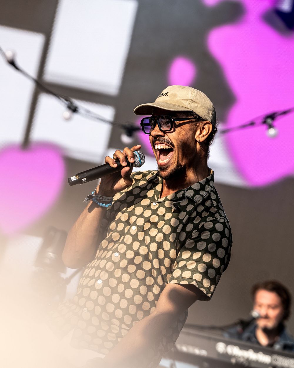 A man in a cap and polka dot shirt sings passionately into a microphone on stage at an outdoor concert.