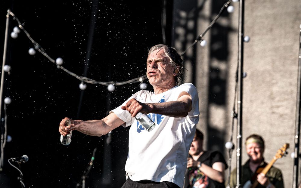 Energetic performer in white t-shirt spraying water on stage at outdoor music festival.