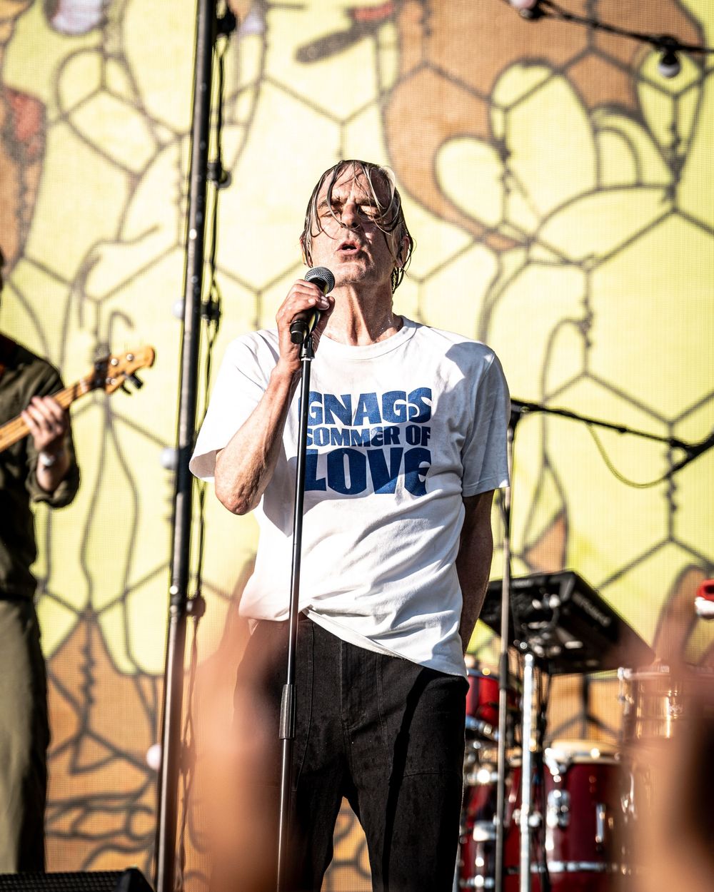 Male singer performing on stage, wearing a white Gnags Summer of Love t-shirt, holding microphone at outdoor concert.