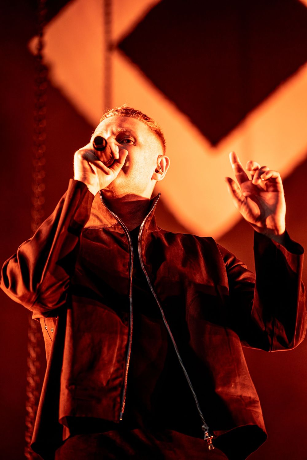 Male performer singing into microphone on stage bathed in dramatic red lighting wearing black jacket.