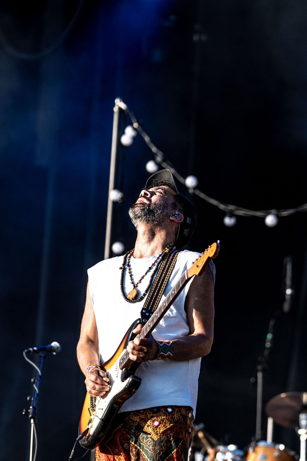 Guitarist in white sleeveless shirt and patterned pants plays electric guitar on stage, head tilted back in musical ecstasy.
