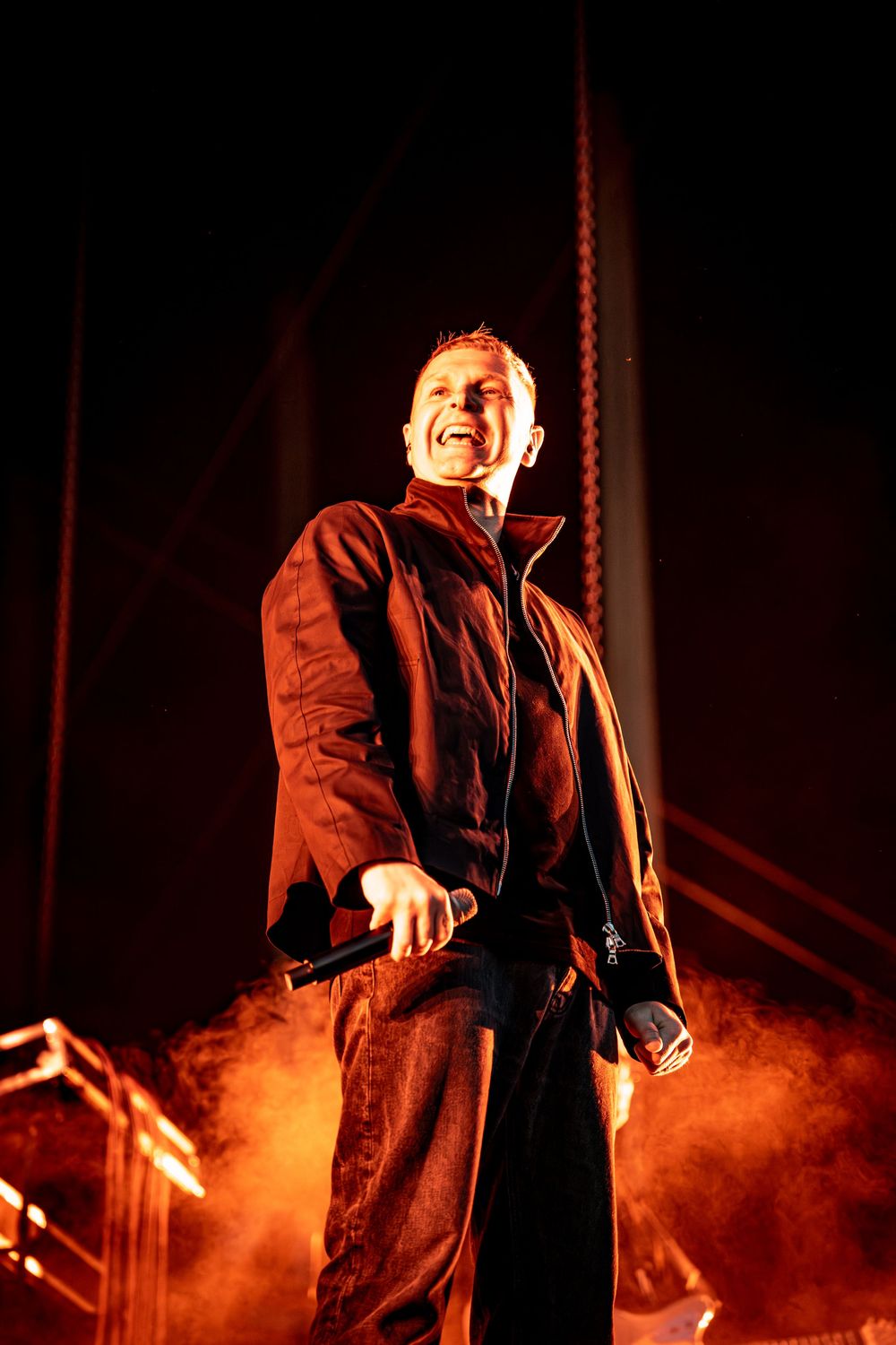 Male performer holding microphone on stage with dramatic red smoke and lighting during a live concert.