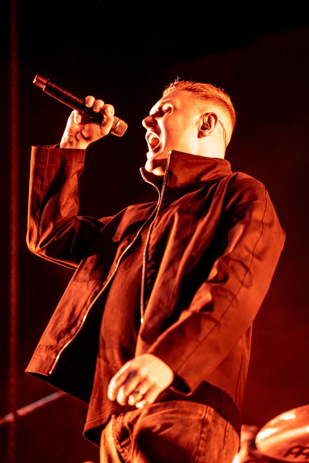 Male singer performing passionately on stage under red lights, holding microphone, wearing black jacket.