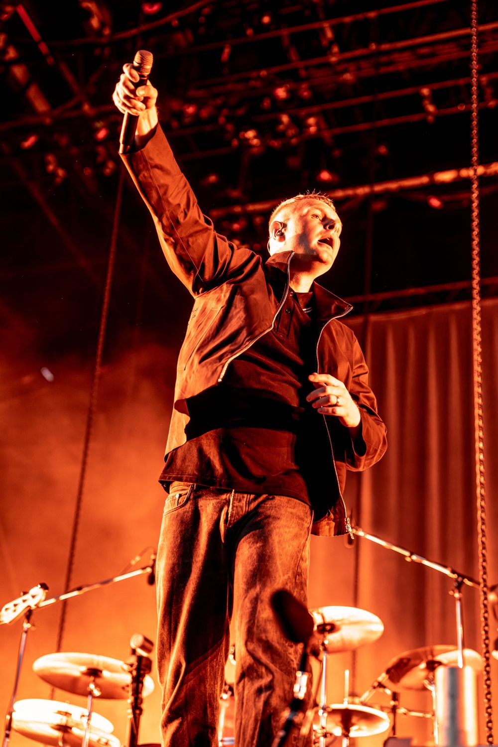 A male performer raises a microphone triumphantly on stage bathed in dramatic red lighting with drums visible behind him.