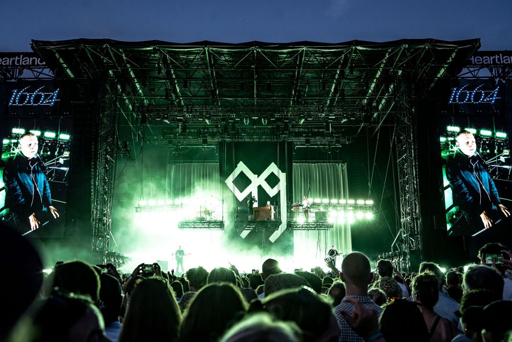 Live concert stage with green lights and large screens showing performers at an outdoor music festival at night.