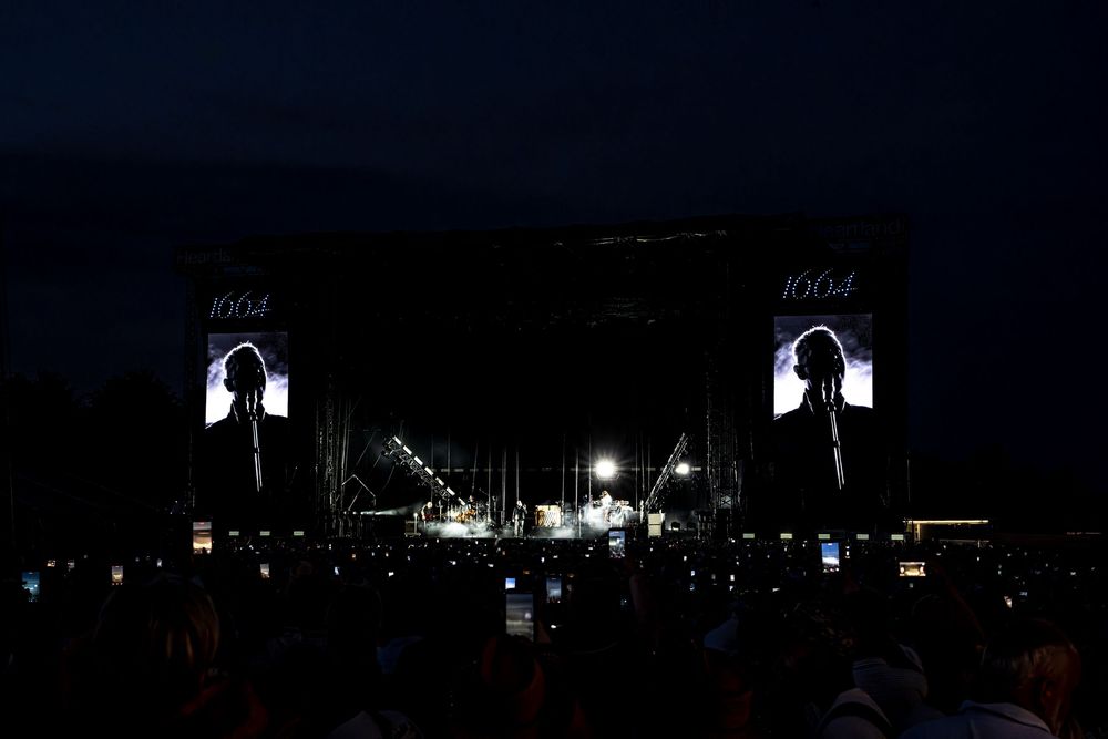 Outdoor concert at night with large screens displaying a performer silhouette above a crowd holding up phones.