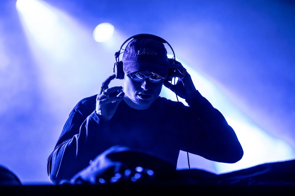 DJ performing live on stage wearing headphones and sunglasses under dramatic blue lighting.
