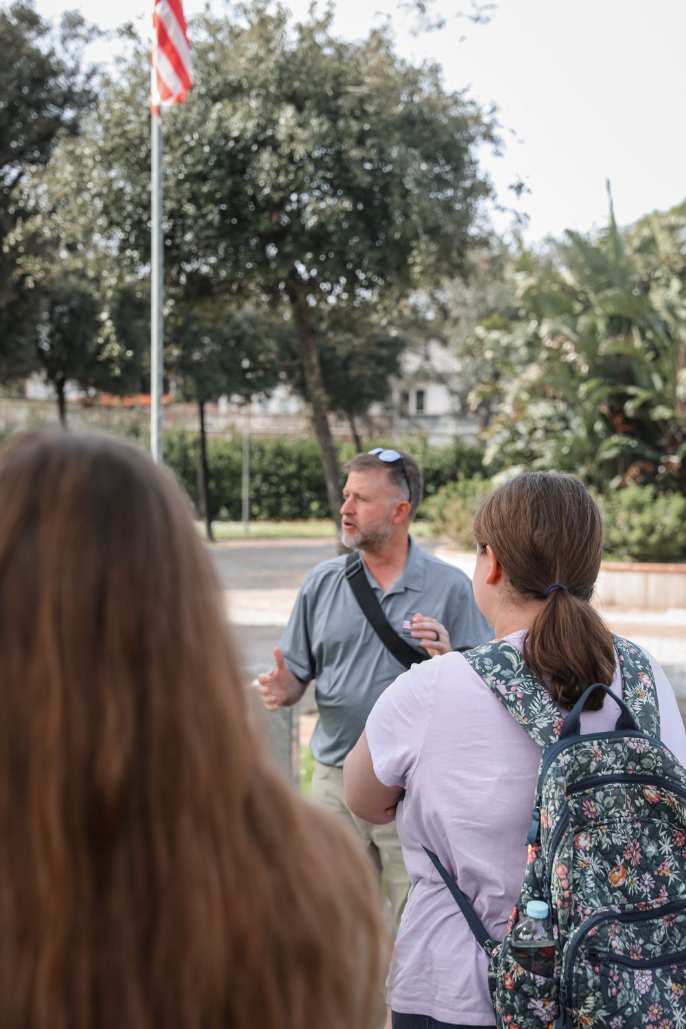A man in a gray shirt leads an outdoor tour group near an American flag on a sunny day.