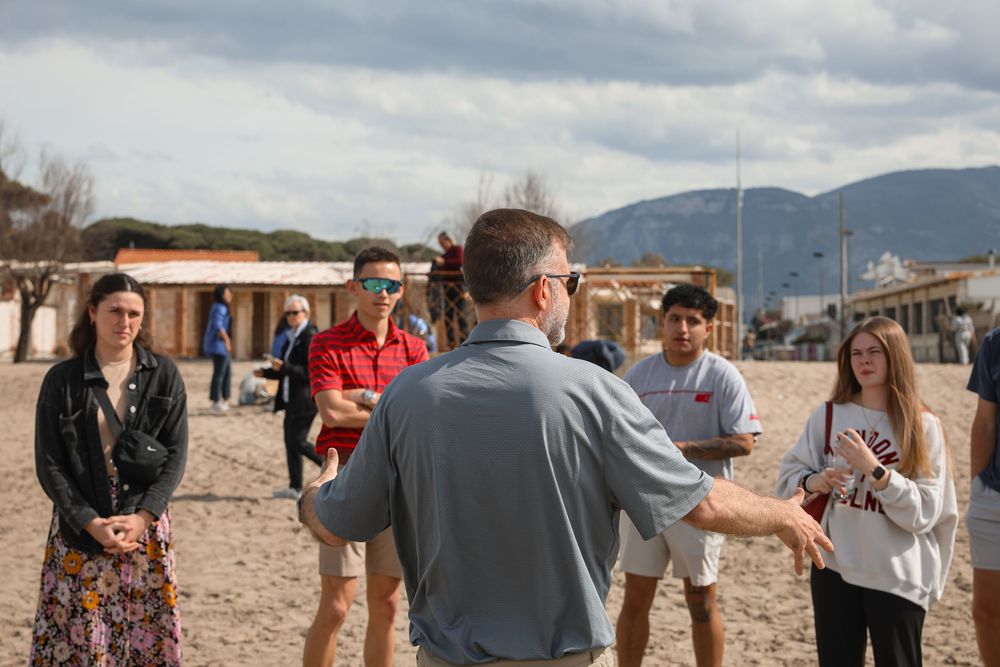 A guide leads a group of students on a sandy beach tour with mountains in the background.