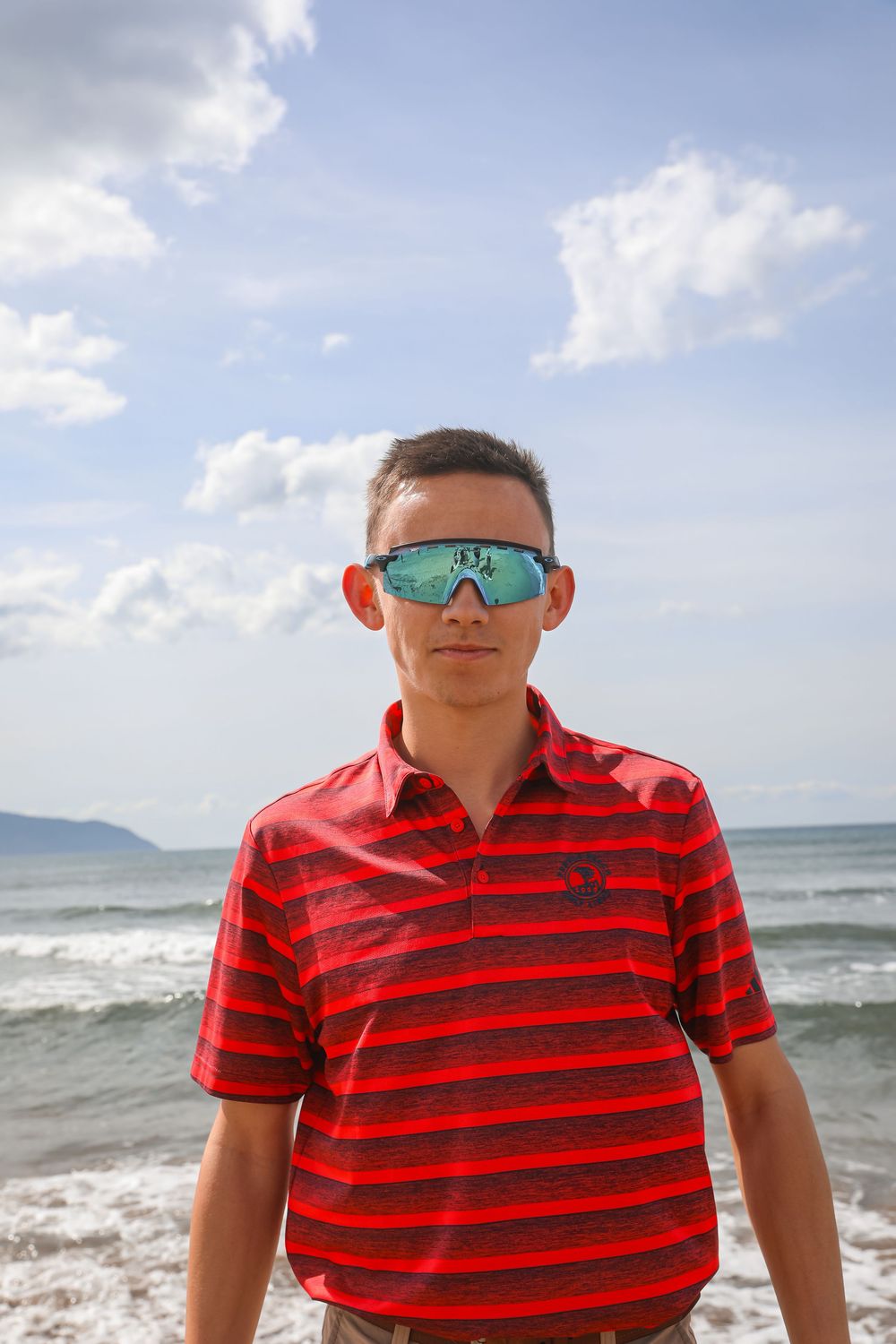 Young man in red striped polo and mirrored sport sunglasses standing on a beach with ocean waves and blue sky.