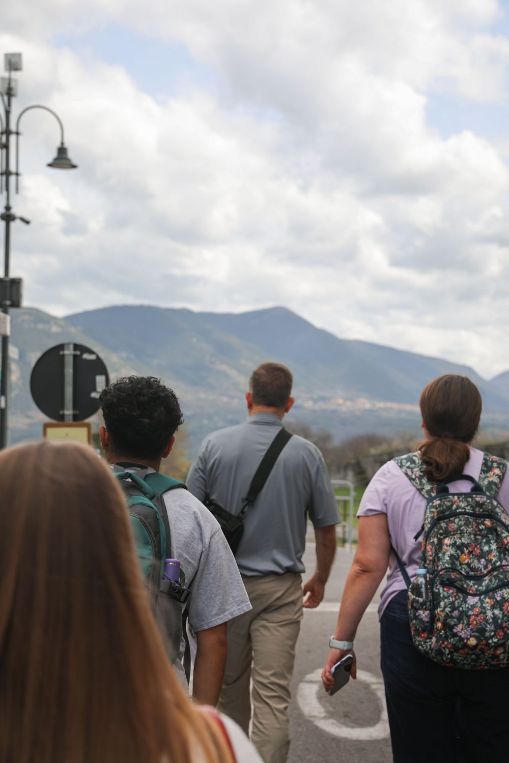 Group of tourists walking toward mountain landscape under cloudy sky, carrying backpacks on a scenic outdoor tour.