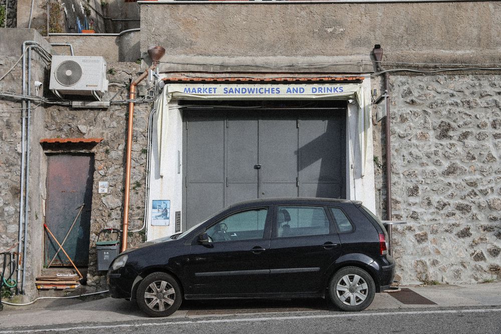 Black hatchback car parked outside a stone building with a sandwich and drinks shop with metal shutters.