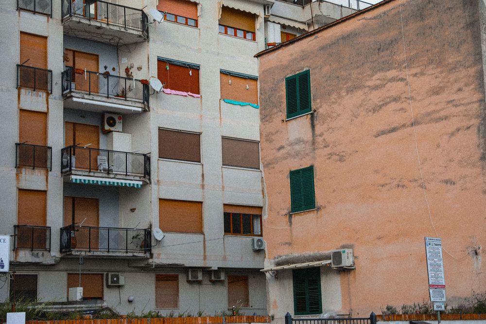 Weathered European apartment buildings with balconies, green shutters, and air conditioning units on an overcast day.