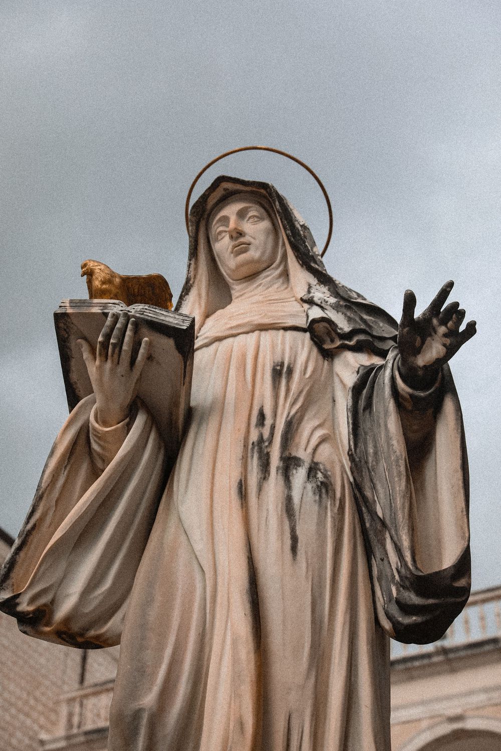Stone statue of a robed saint with halo, holding a book with a small animal, one hand raised, against cloudy sky.