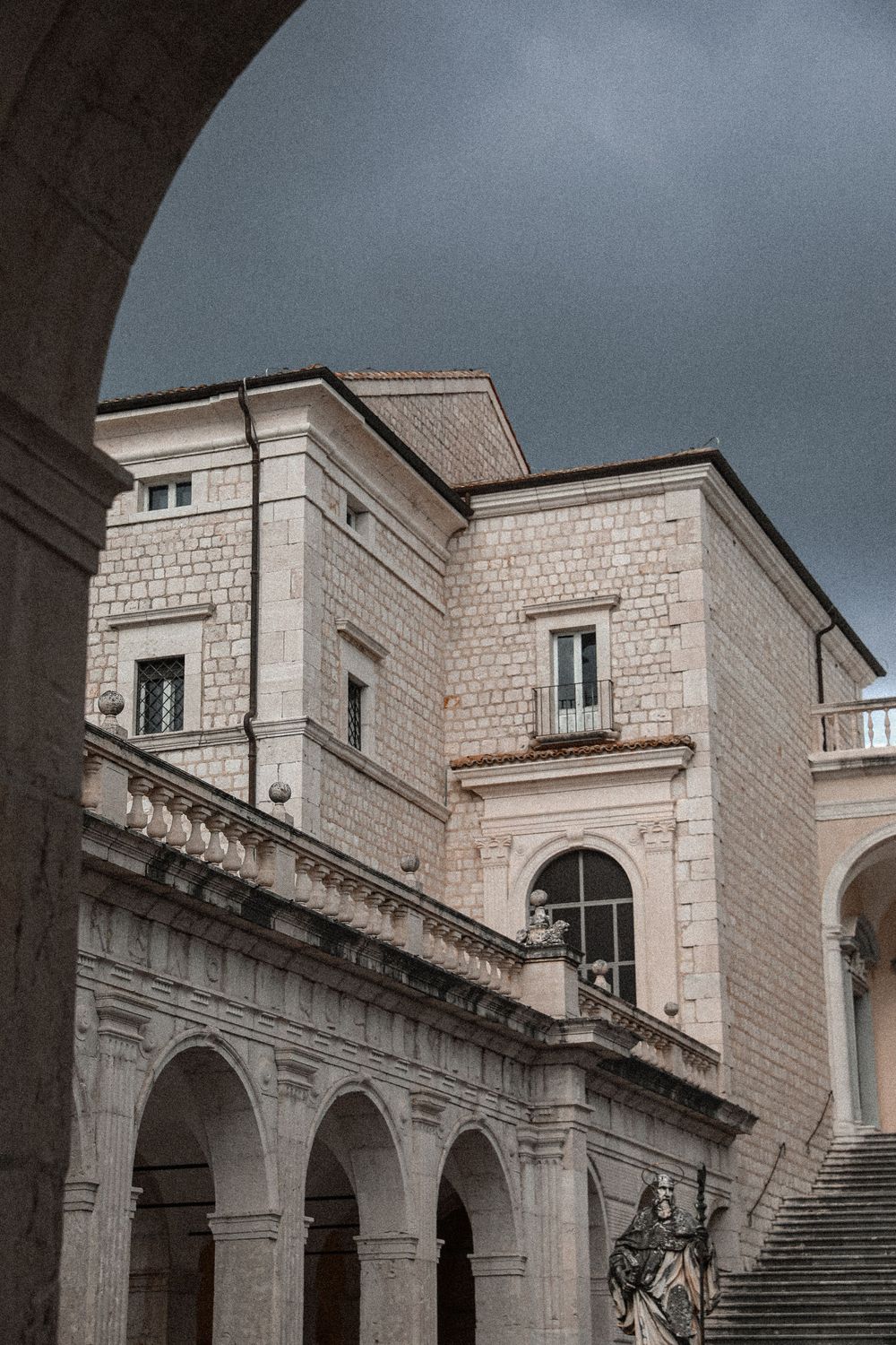 Historic stone building with arched colonnade and statue under dramatic cloudy sky, classical Italian architecture.