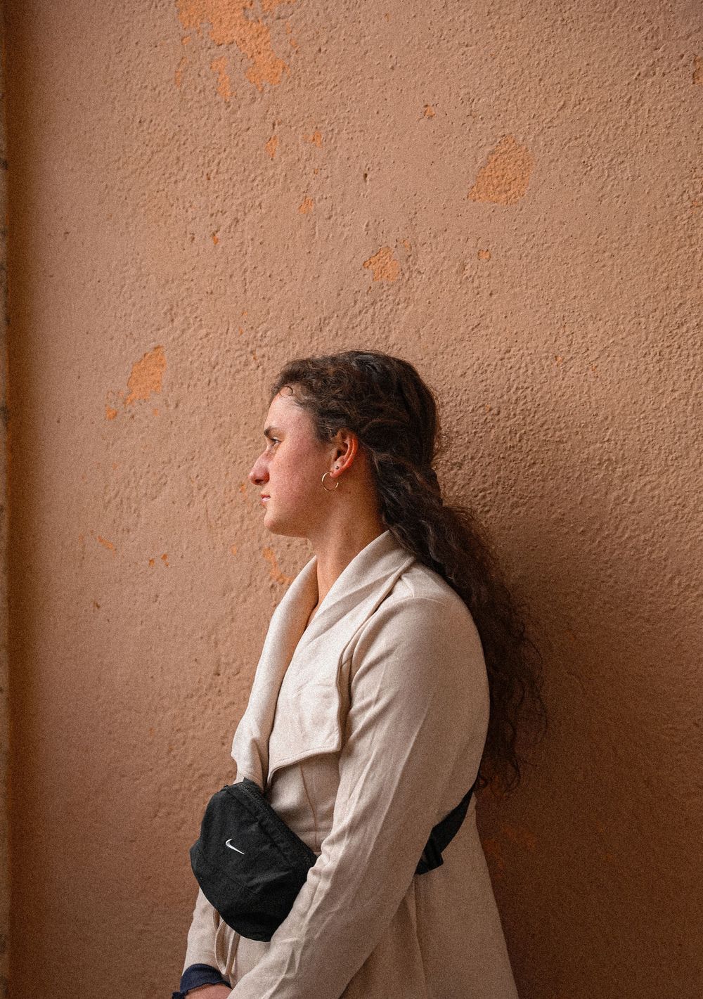 Young woman with curly hair leans against a textured orange wall, wearing a beige coat and Nike bag.
