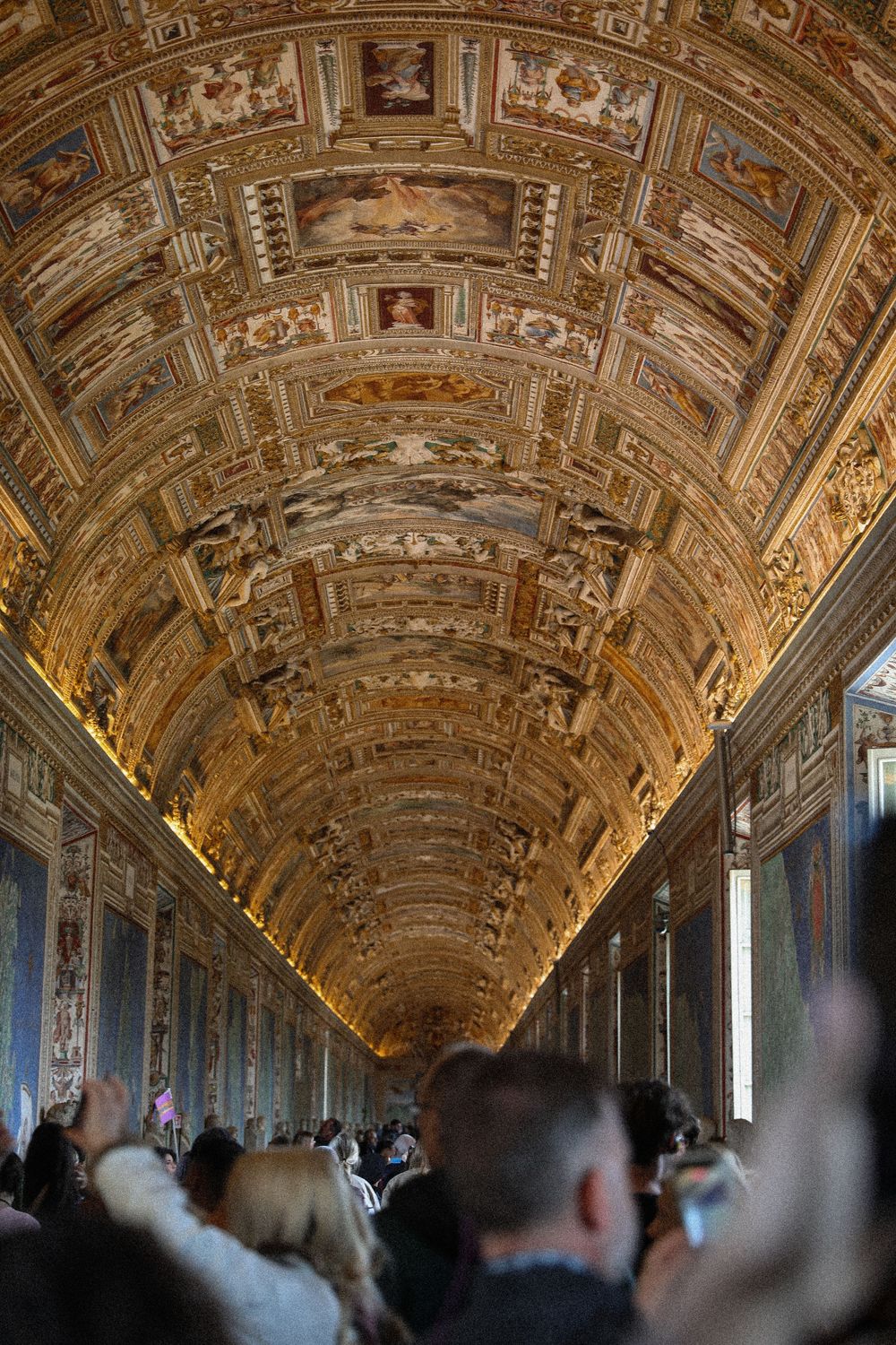 Ornate gilded ceiling of the Vatican Museums Gallery of Maps, crowded with tourists below.