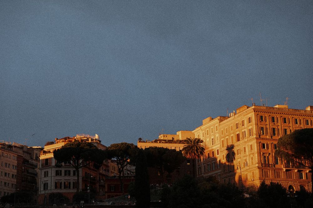 Historic Roman buildings bathed in warm golden sunset light against a dramatic dark blue stormy sky.