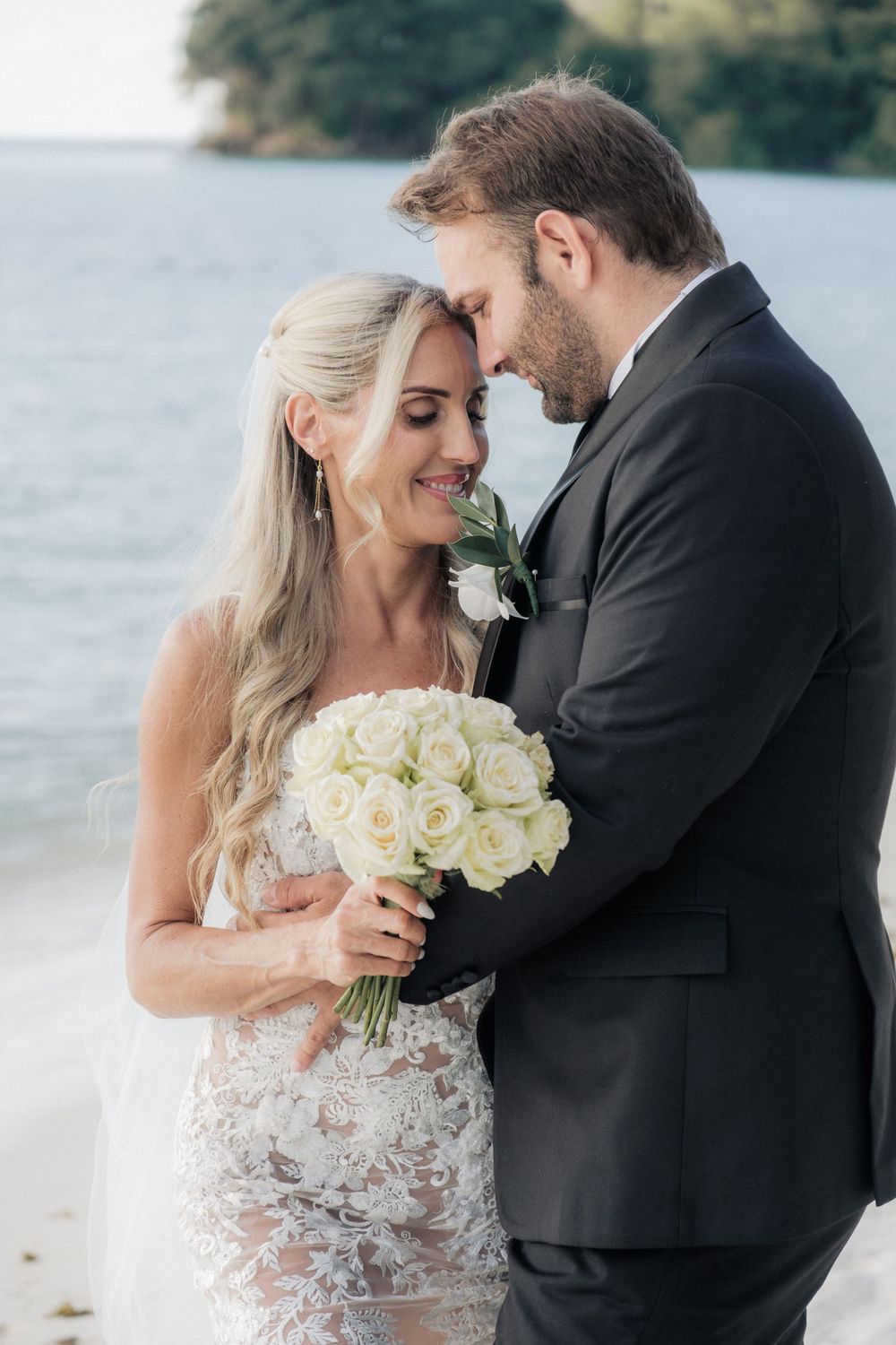 Bride and groom share a romantic moment on a beach, she holds white roses bouquet, both dressed in wedding attire.