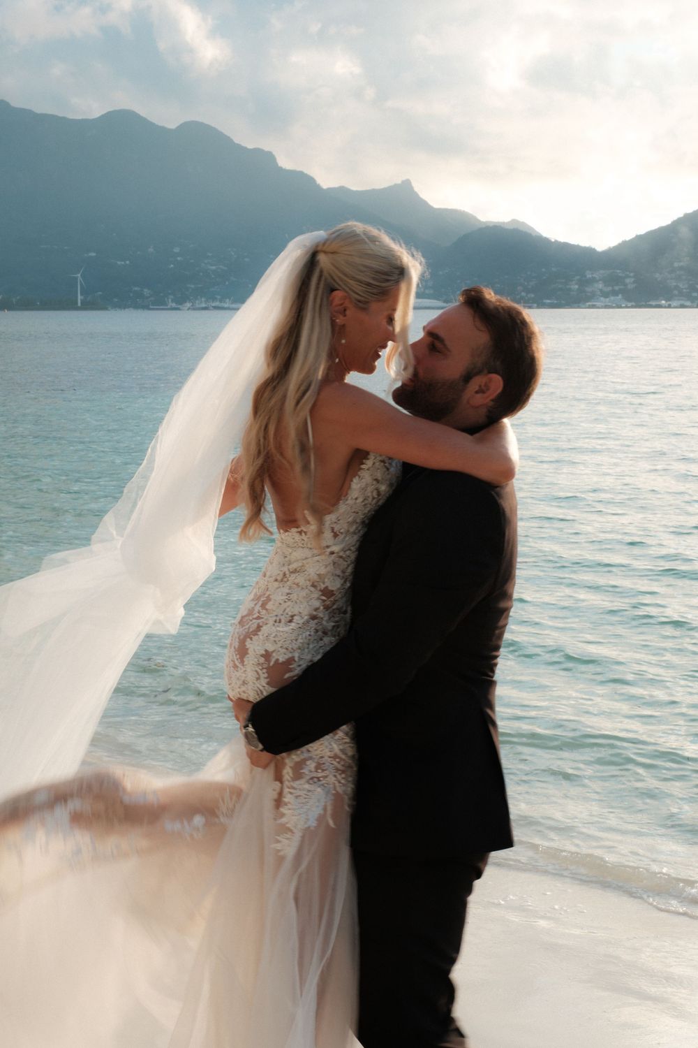 Bride in lace gown and veil embraces groom in black suit on beach with mountains and sea backdrop.
