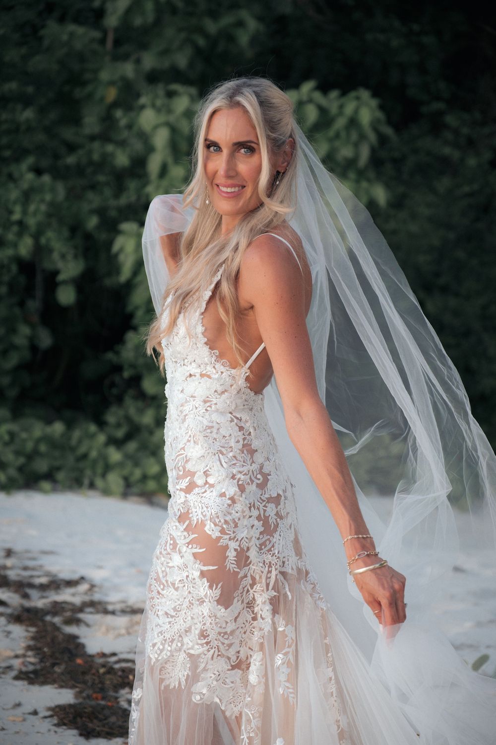 Blonde bride in lace wedding gown and flowing veil poses on sandy beach with lush green trees behind her.