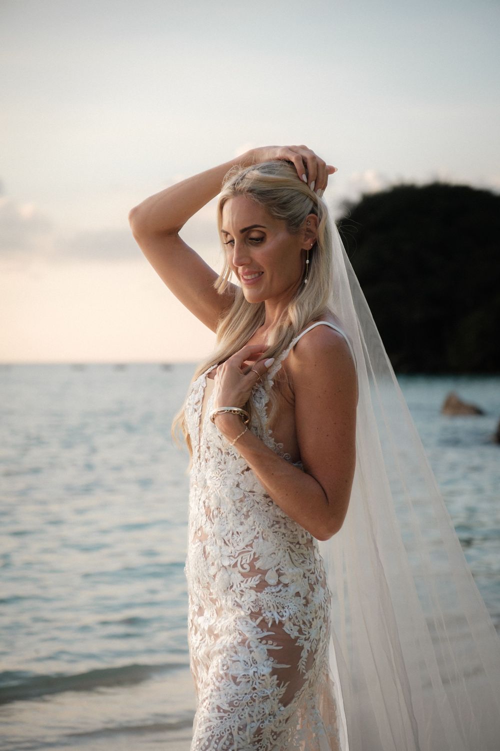 Bride in lace wedding dress and veil poses on beach at sunset, smiling with hands in hair.