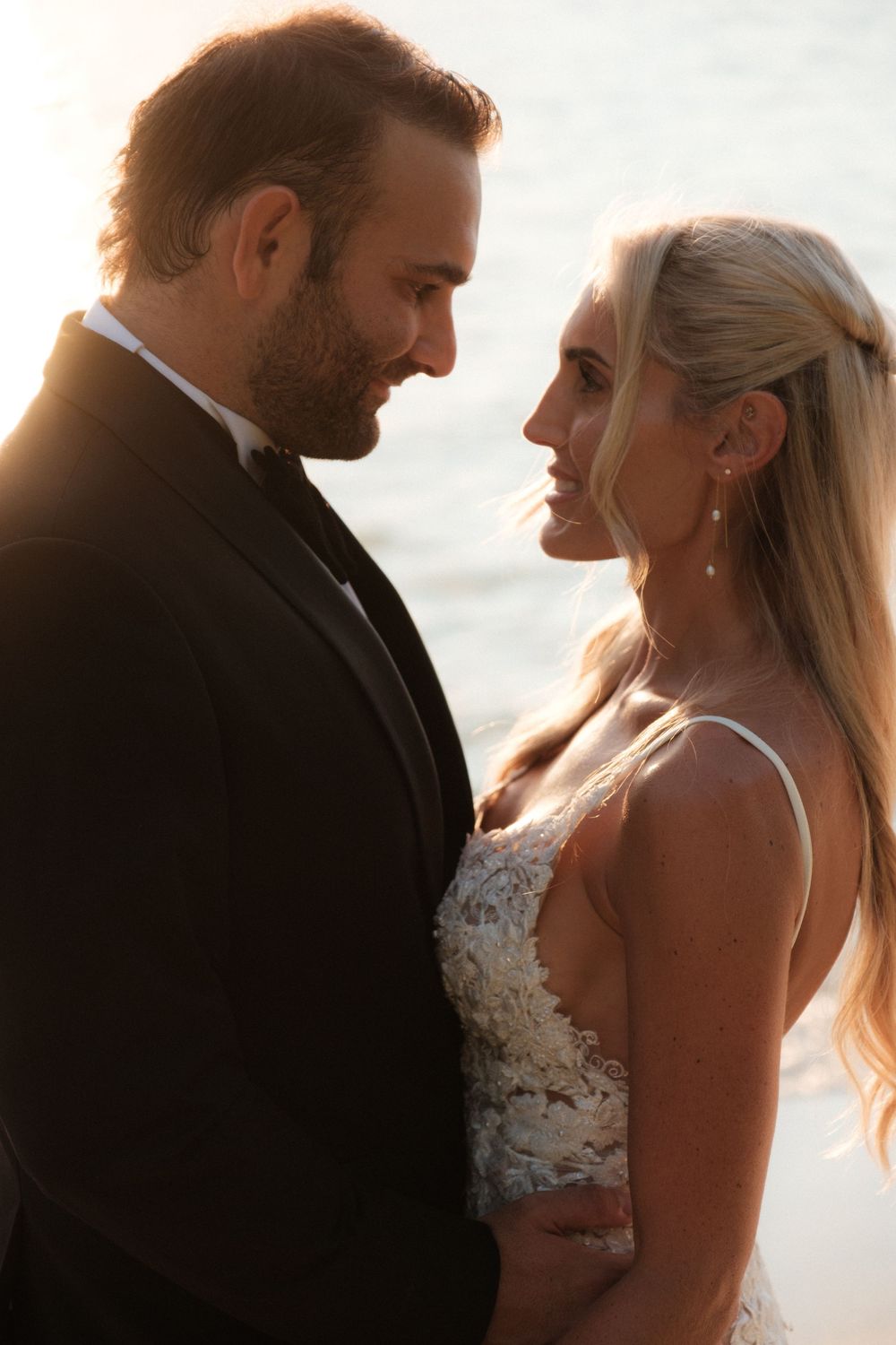 Bride and groom share an intimate moment at golden hour by the water, dressed in wedding attire.