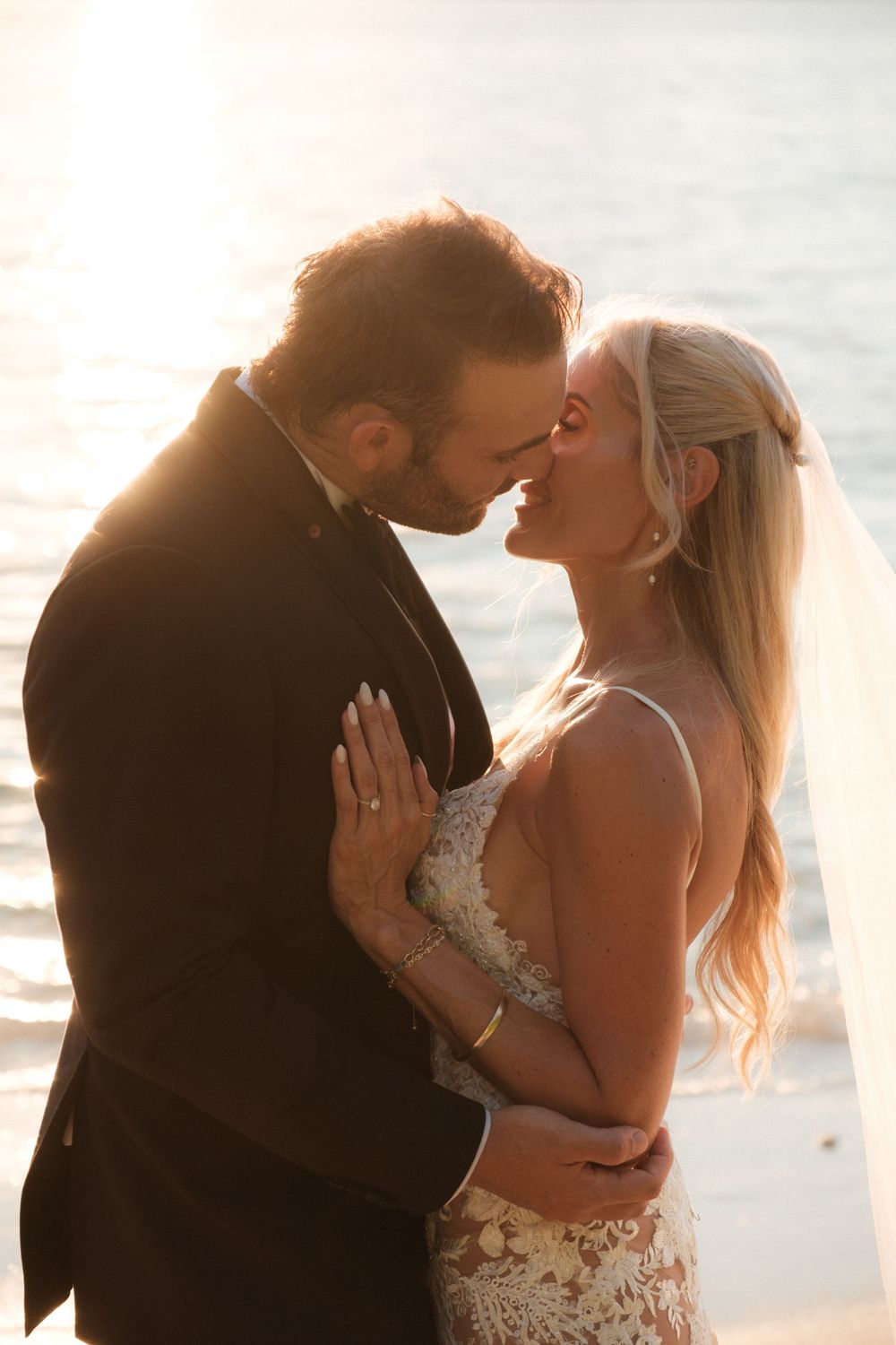 Bride and groom share a romantic kiss at sunset on the beach, golden light glowing behind them.