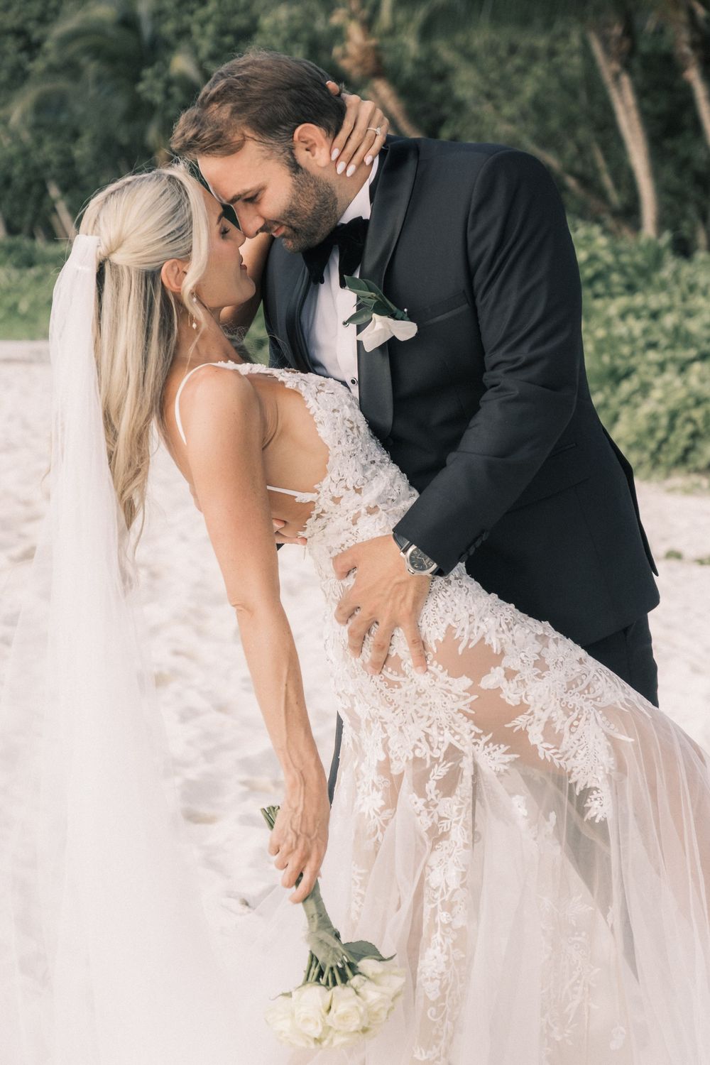 Bride and groom share a romantic moment on a beach, with lace wedding gown, veil, and white bouquet.