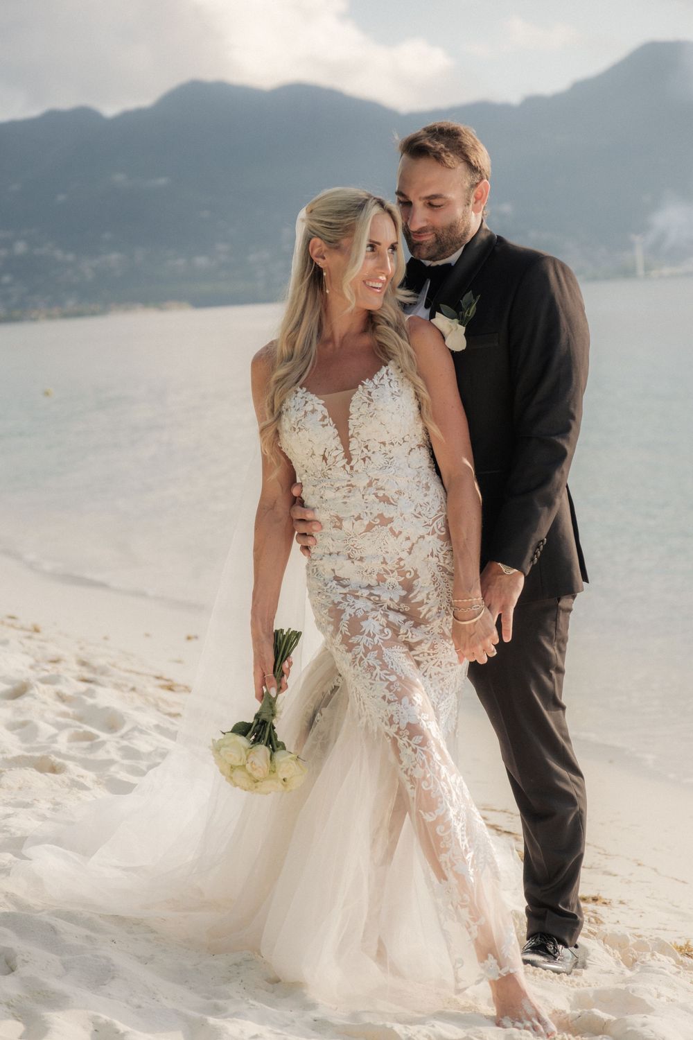 Bride in lace gown holding white bouquet stands with groom in black suit on sandy beach with mountains behind.
