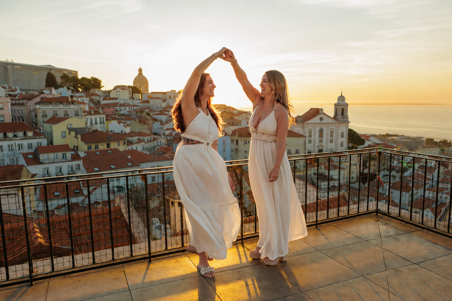 Two women in white dresses dancing on a rooftop terrace at sunset with Lisbon cityscape in background.