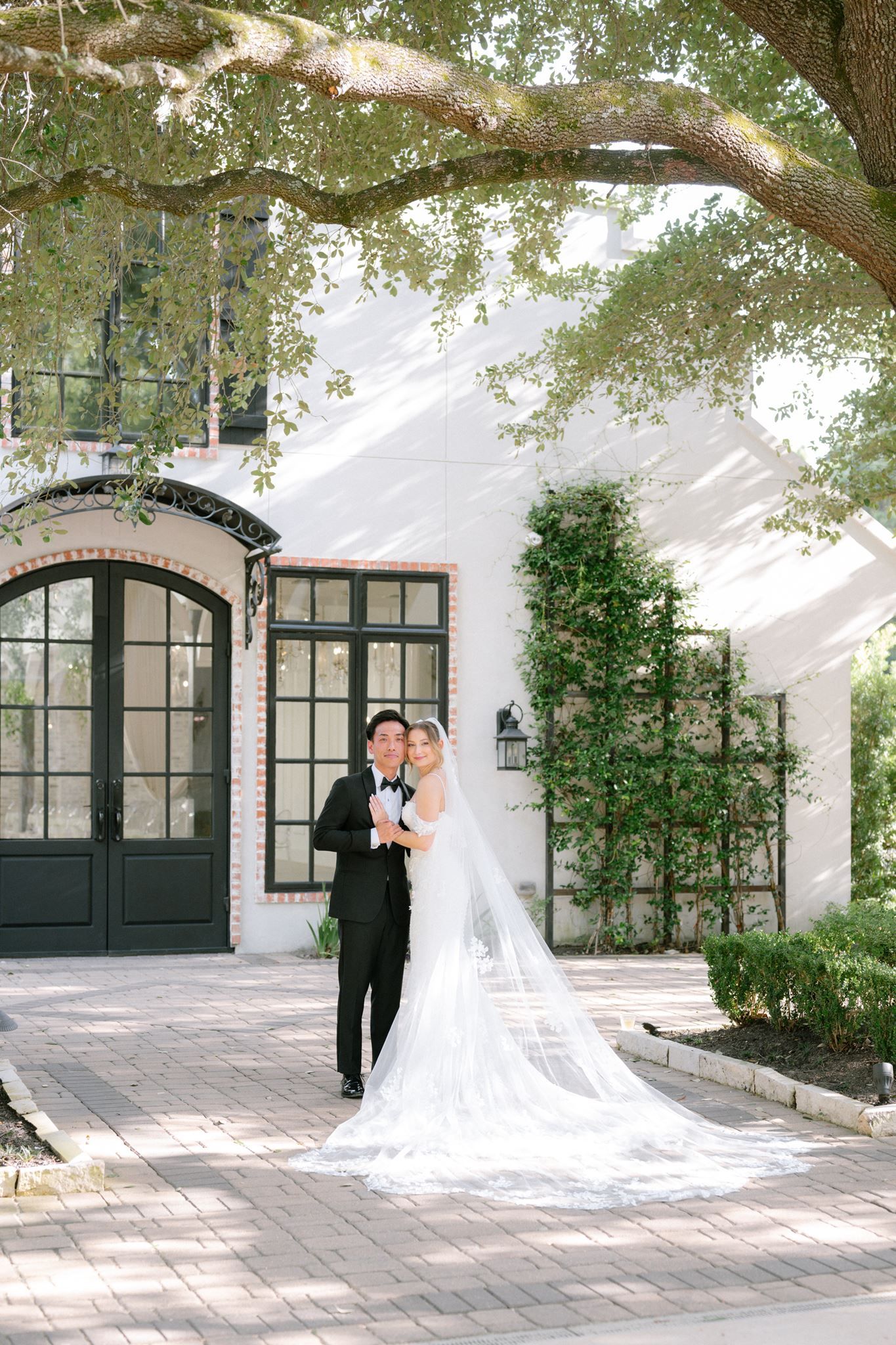 Bride and groom pose together in front of a white brick venue with arched doors and lush greenery on their wedding day.