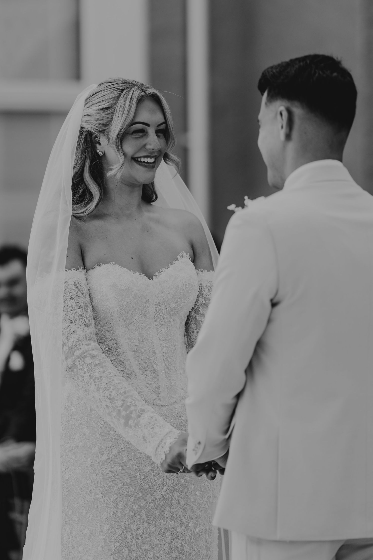 Black and white photo of a smiling bride in lace gown holding hands with groom in white suit at wedding ceremony.