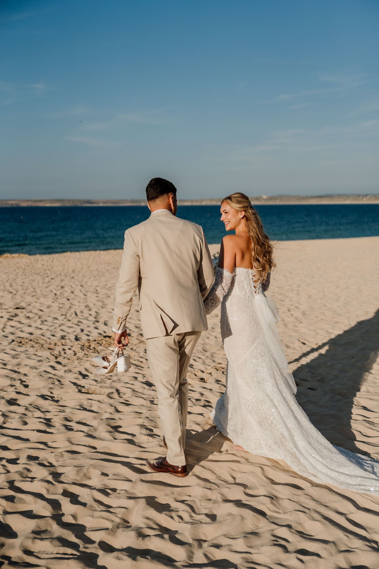 Bride and groom walking hand in hand on sandy beach, bride in lace gown, groom in tan suit, ocean backdrop.
