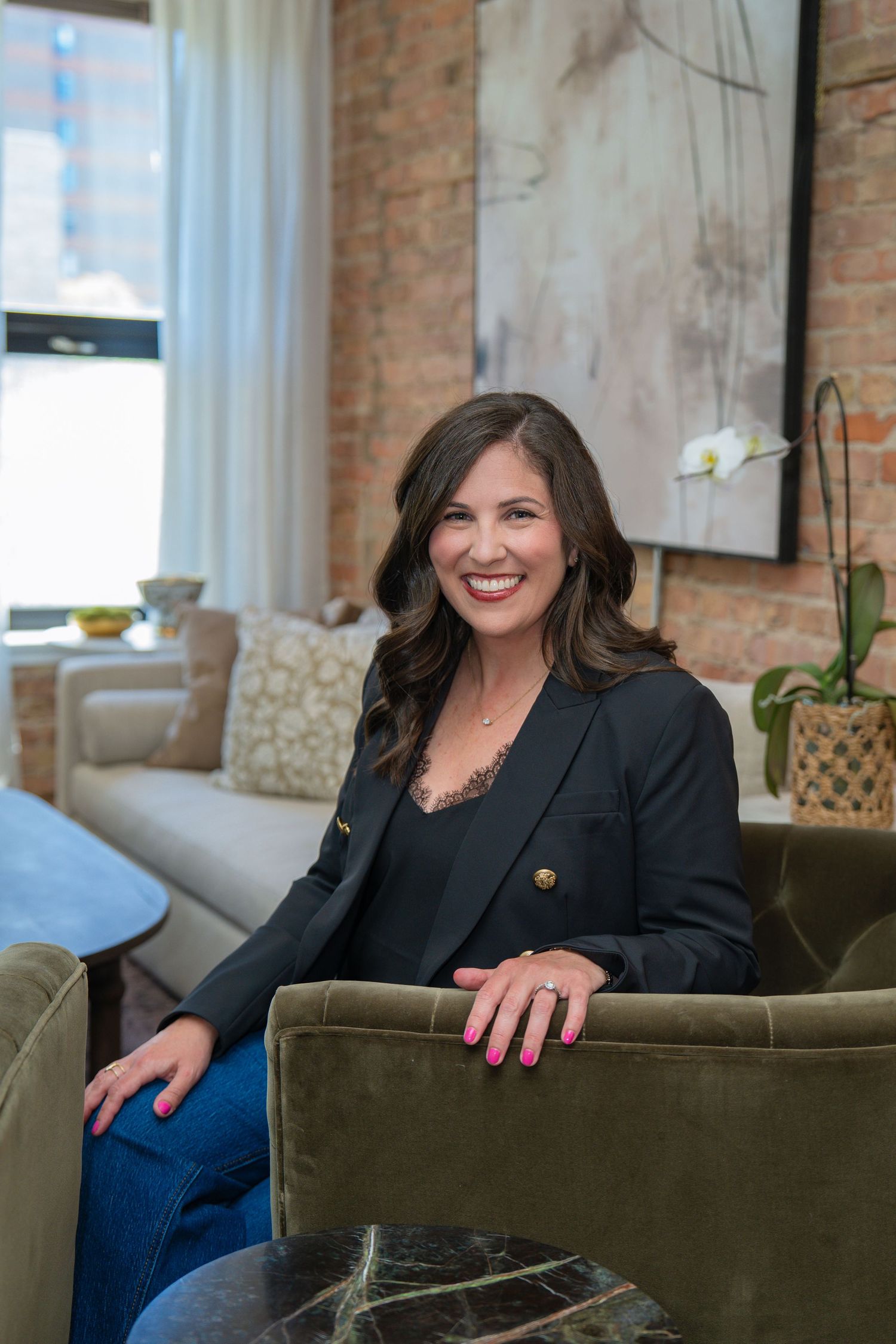 Smiling woman in black blazer and jeans sits in green velvet chair in stylish brick-wall interior space.
