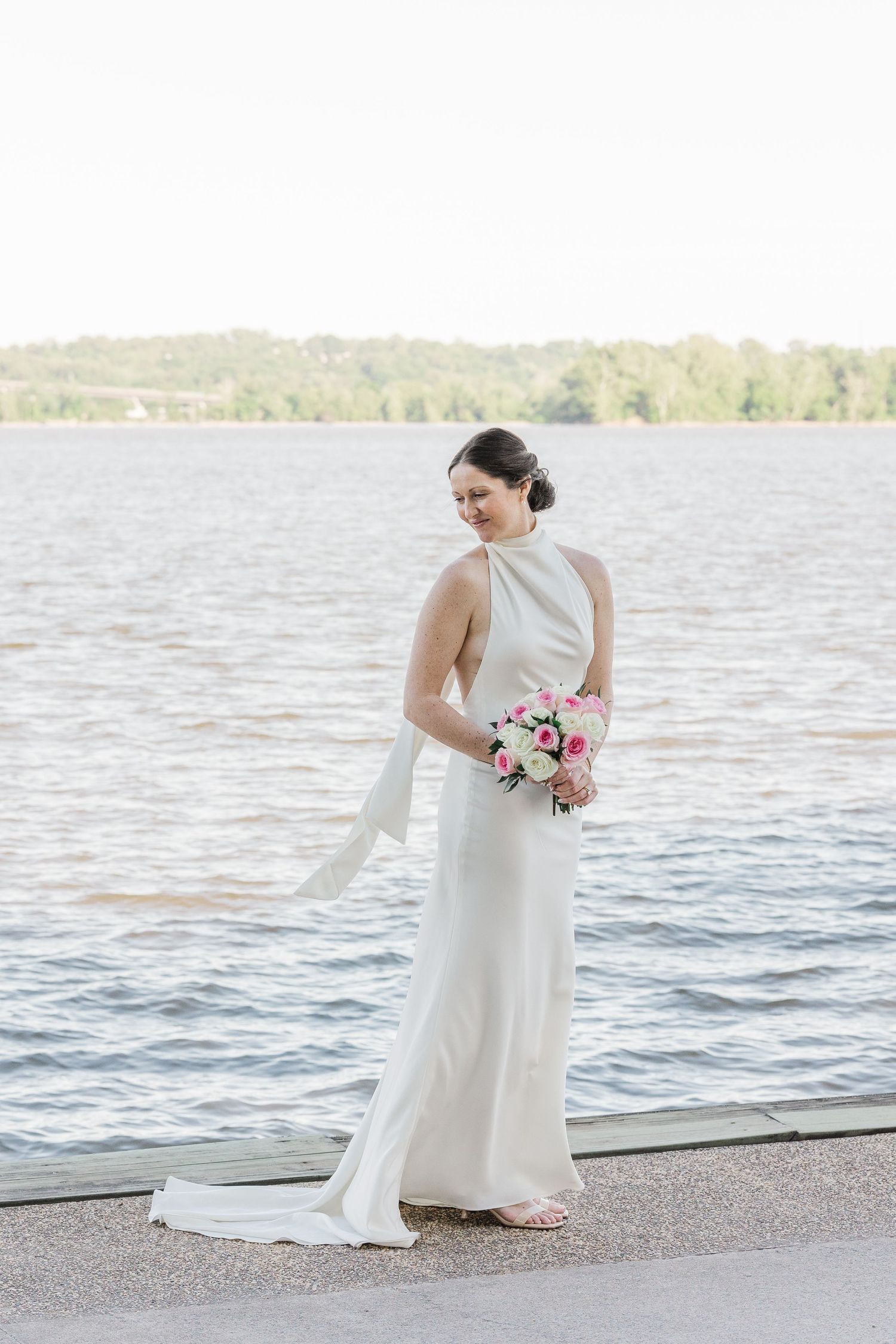 Bride in elegant white halter gown holds pink bouquet by waterfront, her veil flowing gracefully in the breeze.
