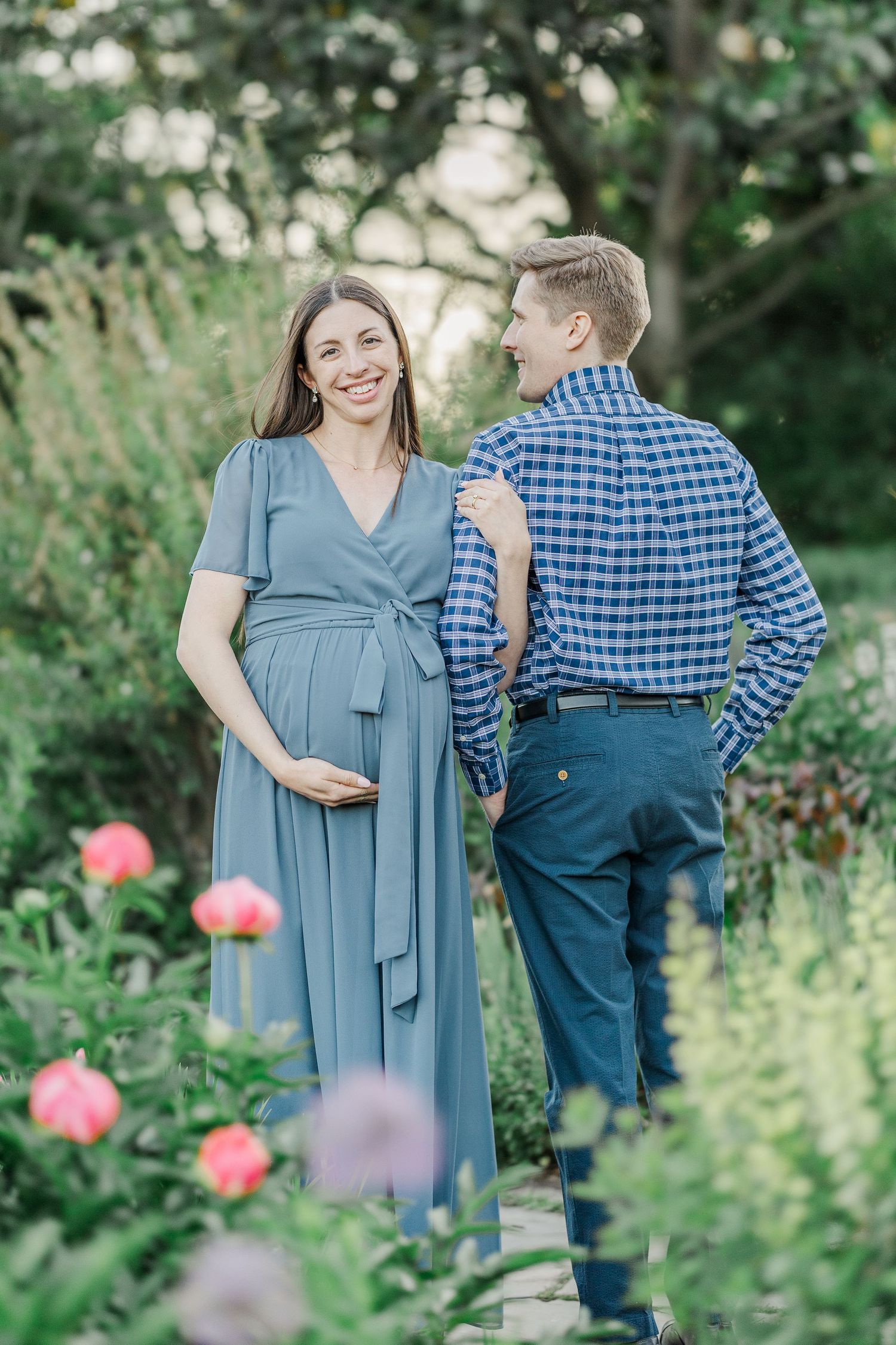 Pregnant woman in blue dress smiles at partner in garden surrounded by blooming pink flowers.