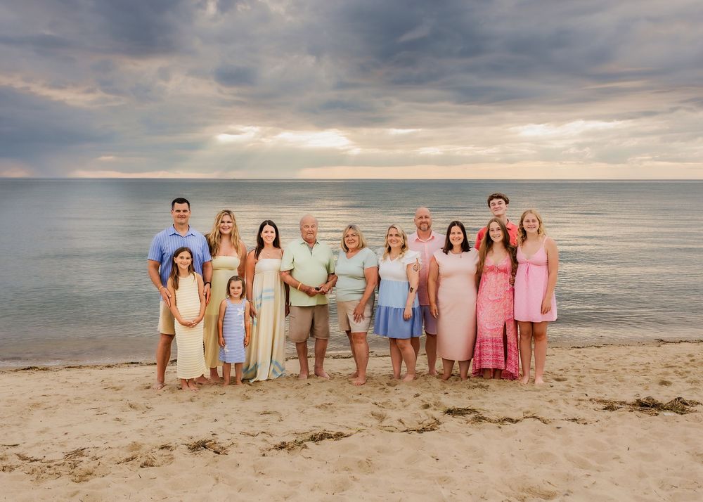 Large extended family poses together on sandy beach at sunset with dramatic cloudy sky backdrop.