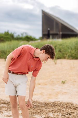 Teen boy in coral polo shirt and khaki shorts bends down on sandy beach near dune grass.