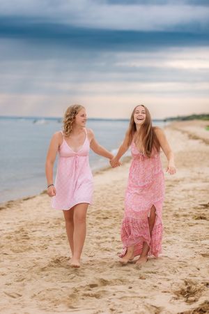 Two young women in pink dresses laugh and hold hands while walking barefoot along sandy beach.