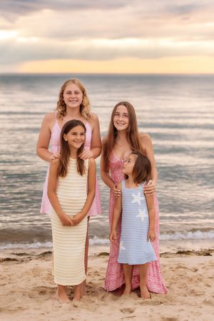 Four sisters pose together on beach at sunset wearing coordinating dresses in pastel colors.