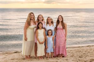 Group of six females including mother and daughters pose together smiling on beach at golden sunset.