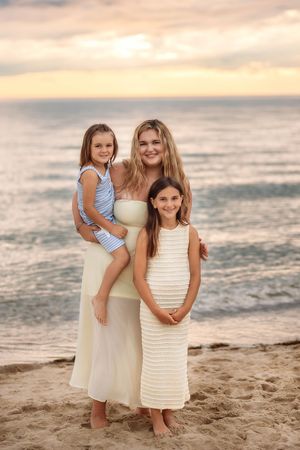 A mother holds her young daughter while standing with her older daughter on a sandy beach at sunset.