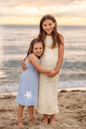 Two young sisters embrace and smile together on a sandy beach during a beautiful sunset.