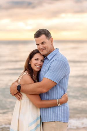 A smiling couple embraces romantically on a sandy beach with a colorful sunset sky behind them.